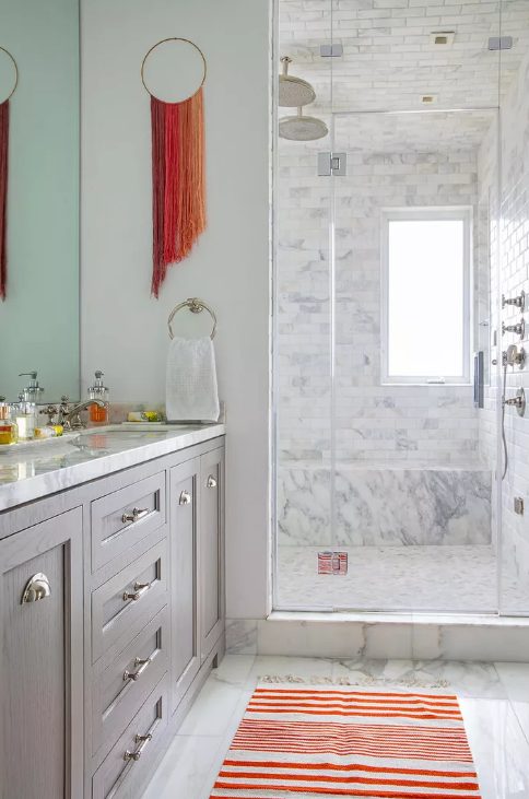a stylish neutral bathroom clad with white marble tiles, with a grey vanity, a bold striped coquelicot rug and a matching hanging with long fringe