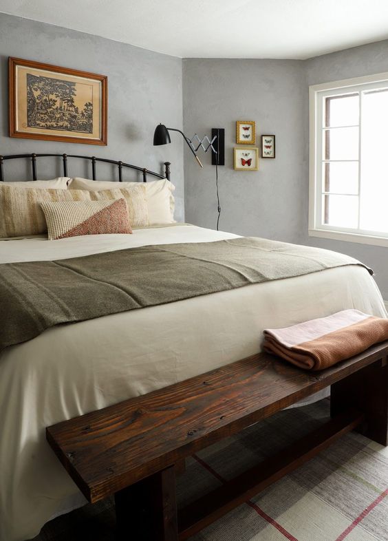 a vintage-inspired bedroom with grey limewashed walls, a black forged bed with neutral bedding, a small gallery wall and a stained wooden bench