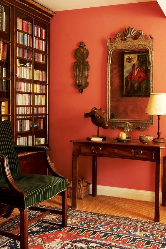 a vintage reading space with a coquelicot wall, heavy dark-stained furniture, a bookcase, a mirror and vintage artwork