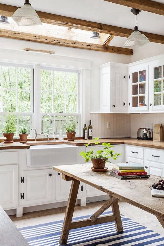 a welcoming neutral farmhouse kitchen with rough wooden beams, white cabinets, butcherblock countertops and a rough wood dining table