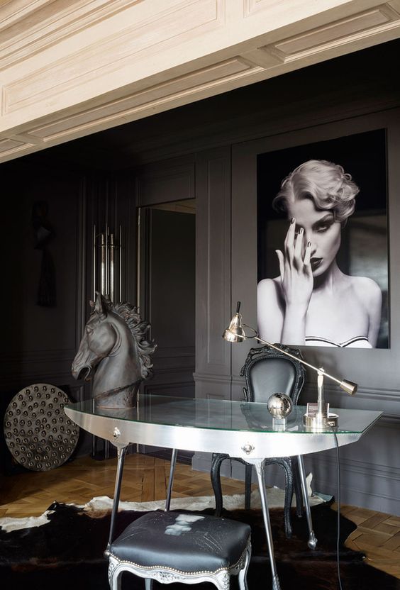 a whimsical and sophisticated space with slate grey walls, a glass and metal desk, vintage chairs and bold artworks