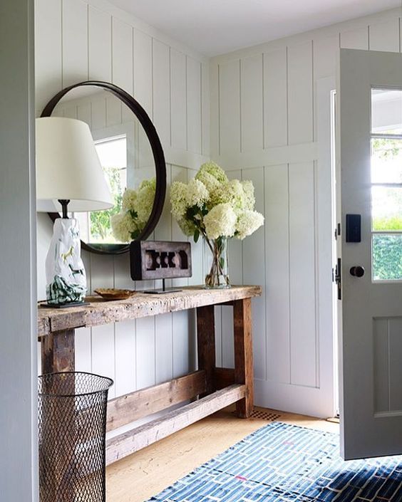 a white planked entryway with a rough wood console table, neutral blooms, a catchy table lamp and a mirror with a black frame