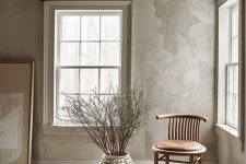 an airy space with grey limewashed walls, a wooden ceiling, a leather and wood chair, branches in a vase