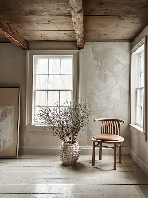 an airy space with grey limewashed walls, a wooden ceiling, a leather and wood chair, branches in a vase