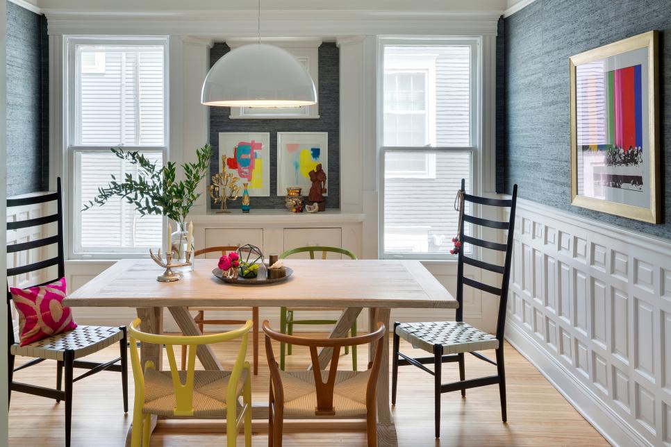 an eclectic dining room with graphite grey grasscloth wallpaper and white wainscoting, a whitewashed table, mismatching chairs and bold art and accessories