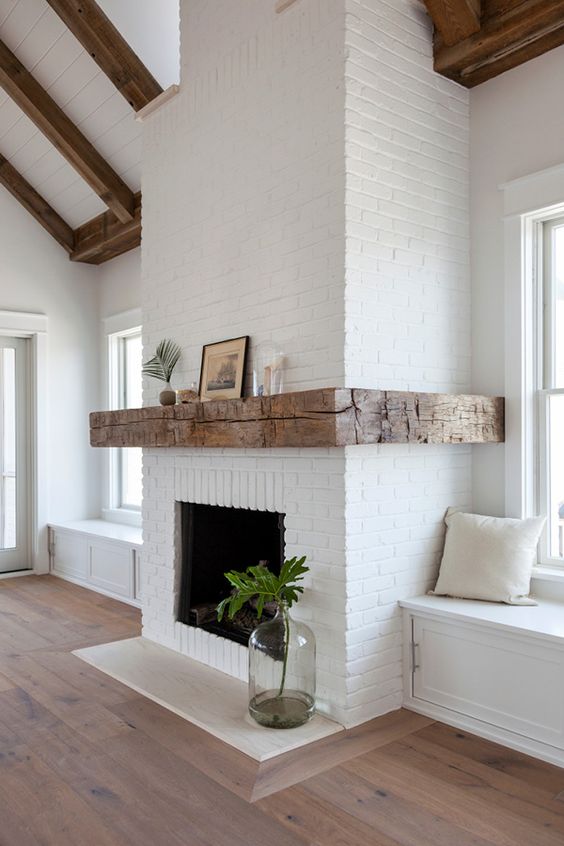 white brick walls and windowsill daybeds, rough wooden beams on the ceiling and a wide rough wood mantel that contrasts the brick