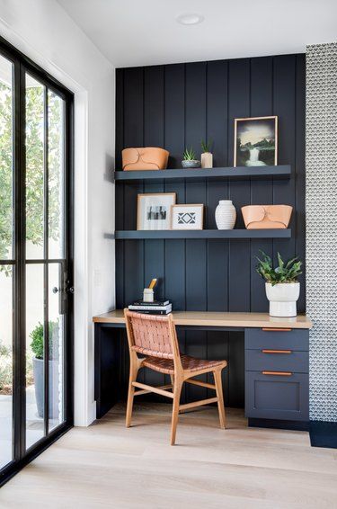 a small navy nook with shiplap, a built-in desk, open shelves, a woven chair and potted plants and leather baskets
