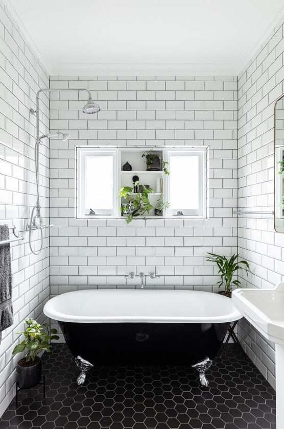 a beautiful black and white bathroom with white subway and black hex tiles, a free standing sink, a black vintage bathtub and a window