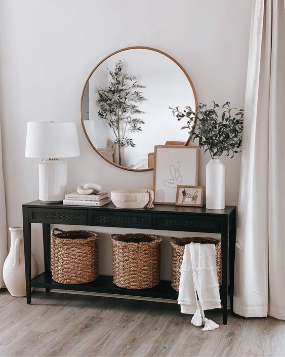 a black wooden console table, three baskets for storage, books, art and a bowl, a white table lamp and greenery branches in a vase