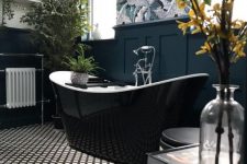 a chic bathroom with a wallpaper accent wall, navy paneling, a glossy black bathtub, printed floor and lots of greenery and blooms