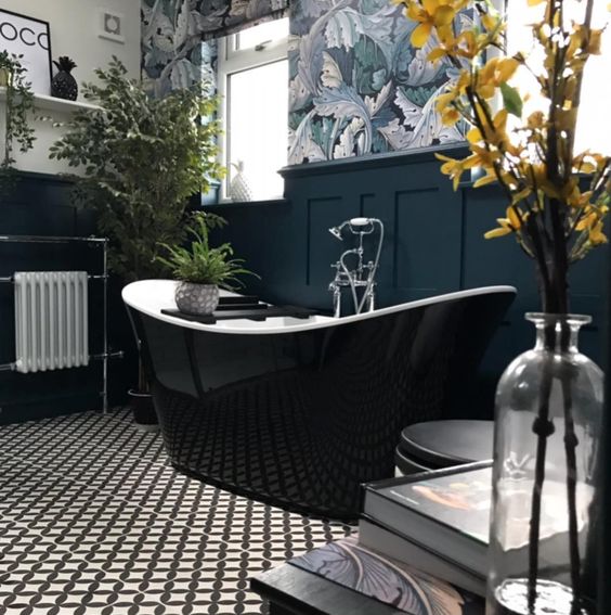 a chic bathroom with a wallpaper accent wall, navy paneling, a glossy black bathtub, printed floor and lots of greenery and blooms