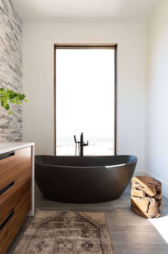 a chic farmhouse bathroom with a faux stone accent wall, a grey tile floor, a light-stained vanity, a black stone bathtub