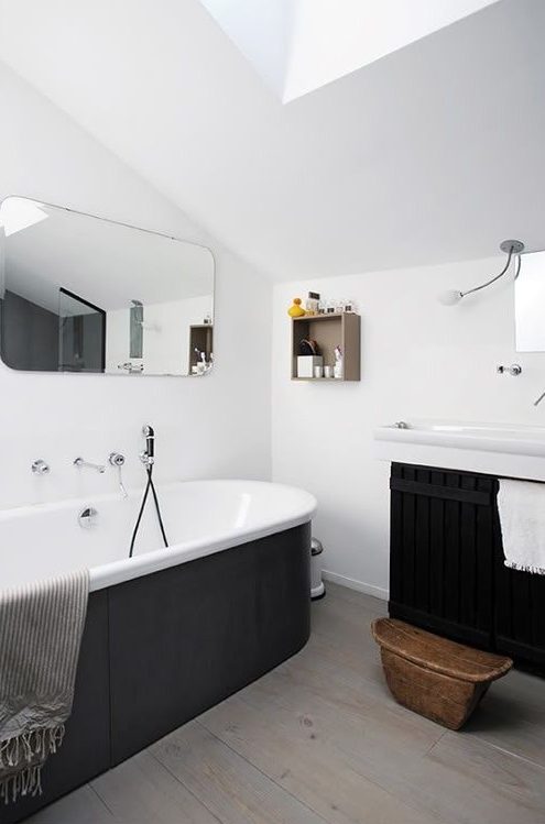 a contemporary black and white bathroom with white walls and laminate on the floor, a tub clad with black panels, a black paneled floating vanity and a rounded mirror