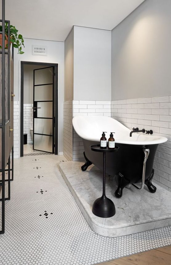 a cool modern bathroom with white subway tiles and penny ones, a black clawfoot tub, a brown vanty and a side table is all chic