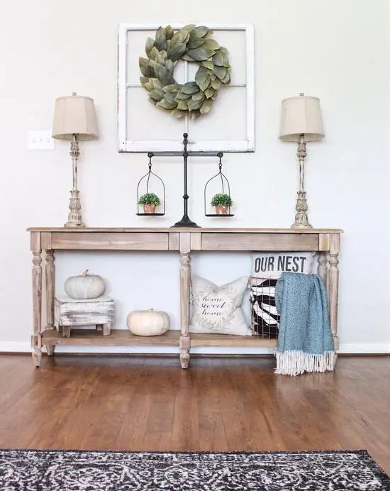 a fall farmhouse console table with white pumpkins, pillows and greenery in pots, with a vintage window frame and a leaf wreath on it