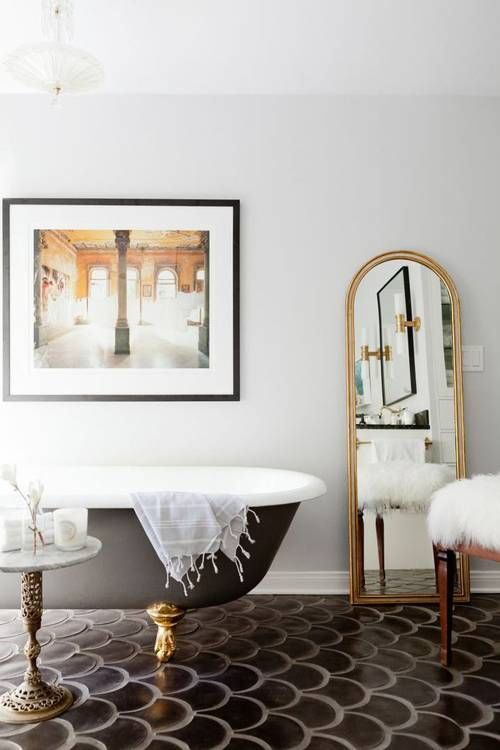 a lovely bathroom with white walls and black scallop tiles on the floor, a black clawfoot bahtub and a chic floor mirror plus a lovely print