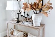 a lovely modern farmhouse console table of wood, baskets with pillows, multiple candles, a chic lamp with a glass base and a vase with leaves