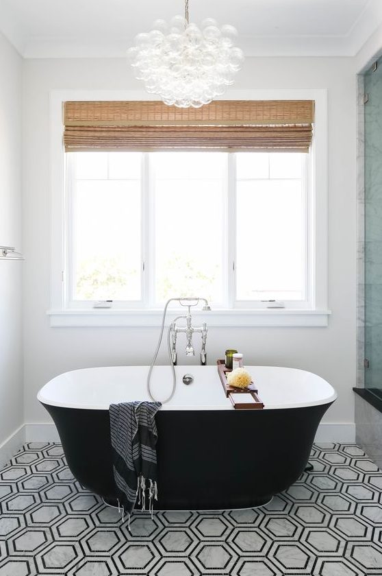 a modern bathroom with black and white hex tiles, a small black tub and a shower space, shades and a bubble chandelier