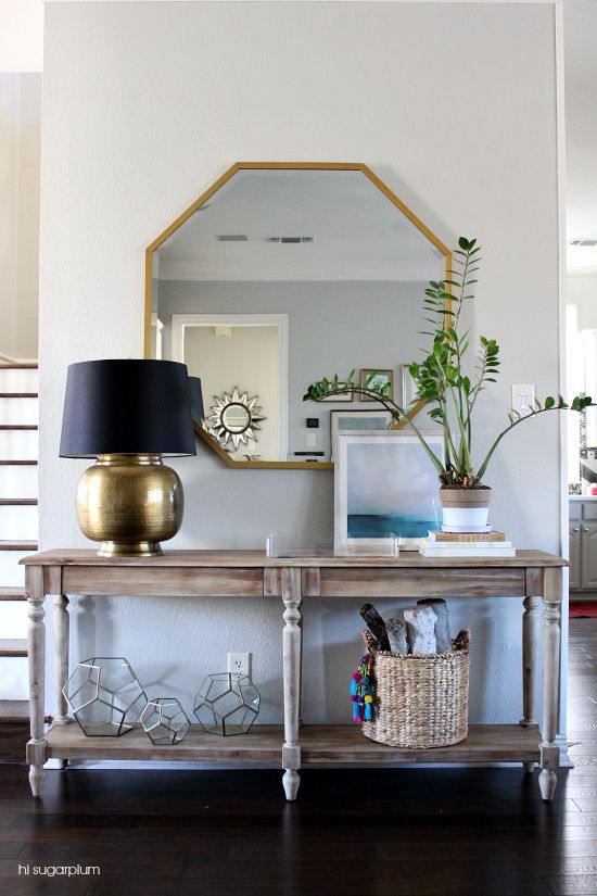 a modern farmhouse console table, faceted candleholders, a brass lamp with a black lampshade, a hexagon mirror and some art