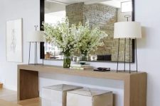 a modern sleek light-stained console table, neutral poufs, matching lamps, an oversized mirror and blooming branches in the vase