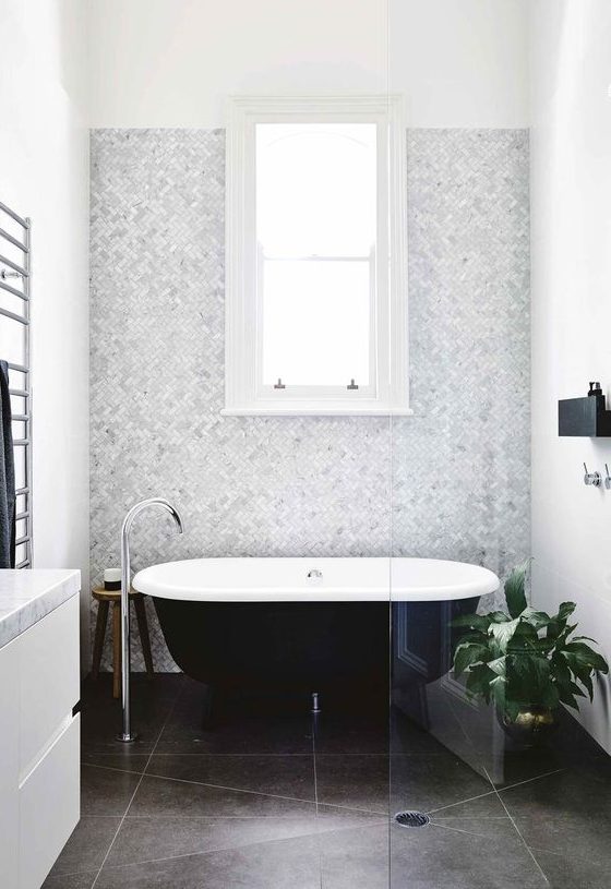 a neutral and chic bathroom with a grey marble accent wall, dark tiles on the floor, a black clawfoot tub, a white vanity and a black mini shelf