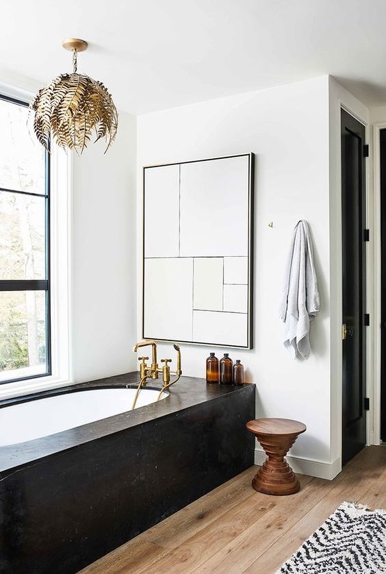 a refined modern bathroom with a tub clad with black stone, a wooden stool, a chic gold leaf chandelier and a large window