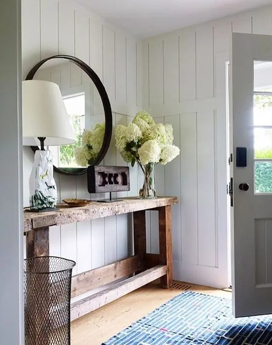 a rough wood console table, a round mirror in a black frame, white hydrangeas, a catchy lamp and a carved wood artwork for a cozy rustic look