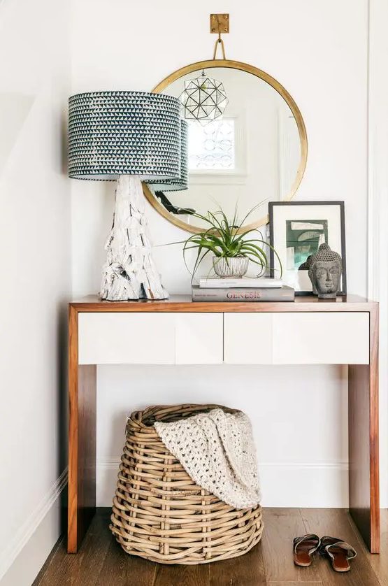 a small mid-century modern console table, a brass frame mirror, a lamp with a driftwood base, a potted plant, some art and a basket