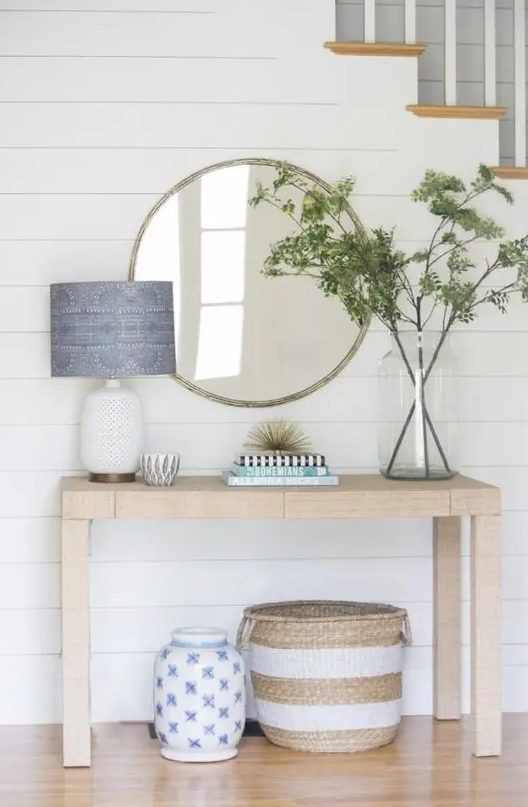 a stylish farmhouse light-stained console table, a round mirror, greenery branches in a large jar, a lamp with a blue printed lampshade and a basket under the table