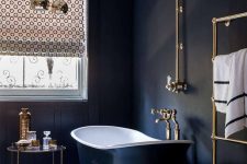 a vintage black bathroom with black walls, a vintage black bathtub, chic brass fixtures and a printed curtain on the window