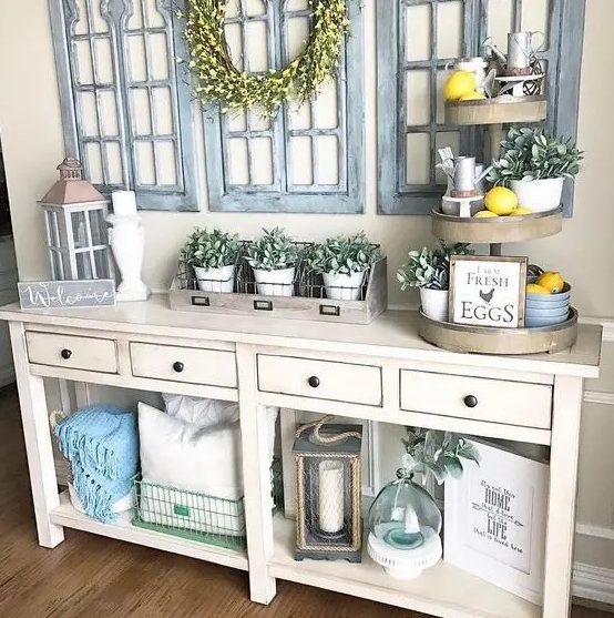 a white farmhouse console table with lots of potted greenery, a faux stand with lemons and greenery, whitewashed window frames, a bright faux flower wreath and blankets and pillows