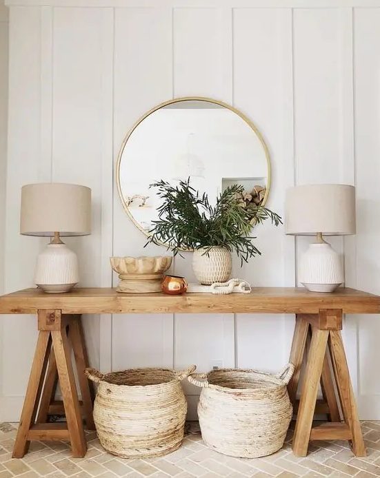 a wooden trestle leg console with a potted plant, some baskets under the console and a couple of elegant lamps plus a round mirror