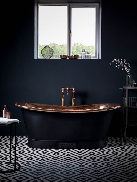 an elegant moody bathroom with matte black walls, a black and copper bathtub, a geometric floor and a window