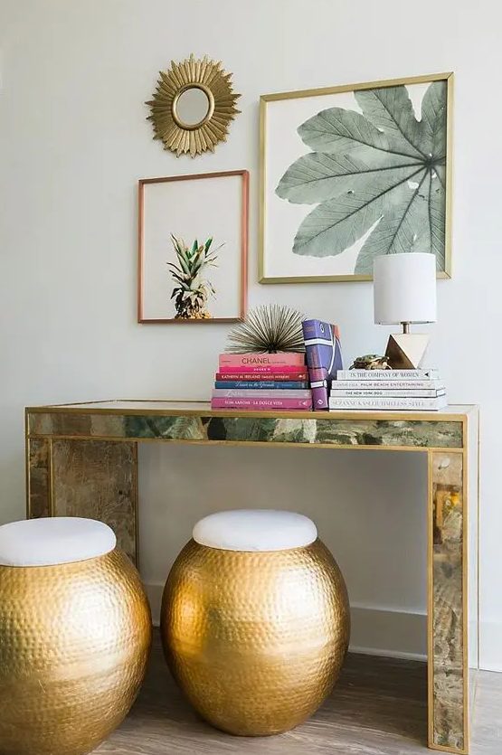 an encrusted console table, colorful books, tropical artworks over the table and hammered metal ottomans