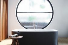 an exquisite contemporary bathroom with a wooden slab accent wall, a wooden floor, a sleek black bathtub and a large frosted glass round window