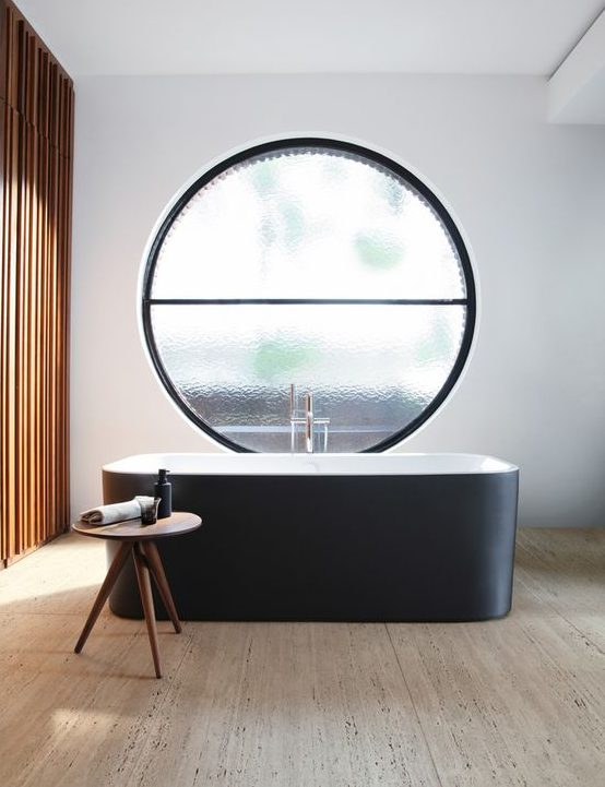 an exquisite contemporary bathroom with a wooden slab accent wall, a wooden floor, a sleek black bathtub and a large frosted glass round window