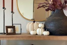 beautiful fall console table styling with faux pumpkins, a black vase with dark foliage, burgundy candles, a mirror in a gold frame and baskets with pillows and pumpkins