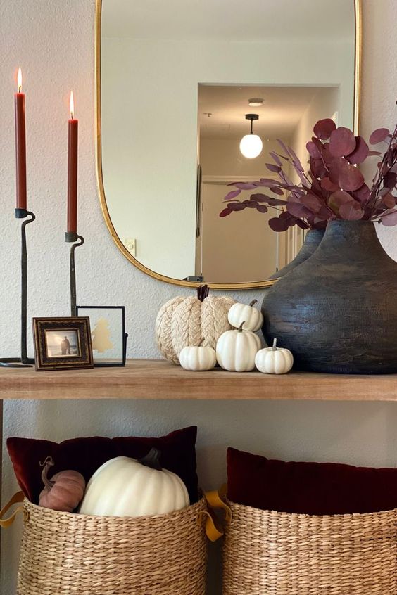 beautiful fall console table styling with faux pumpkins, a black vase with dark foliage, burgundy candles, a mirror in a gold frame and baskets with pillows and pumpkins