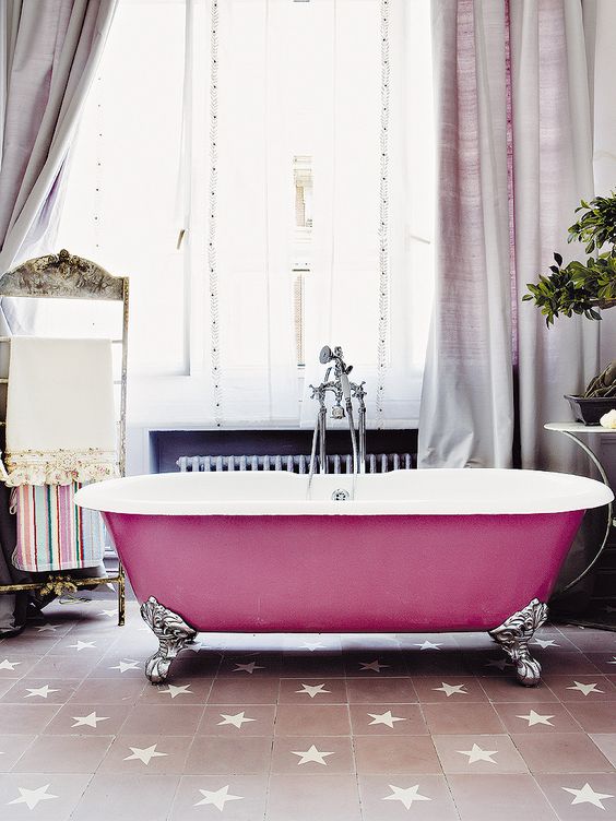 a beautiful bathroom with a blush star print tile floor, a bright pink bathtub, lilac curtains, a stand for towels and potted greenery