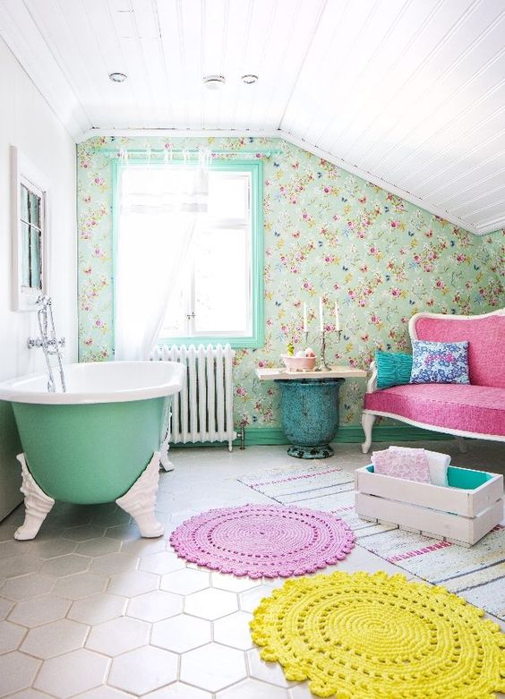 a vintage country bathroom with a floral wallpaper wall, a green bathtub, a pink loveseat, a crate with pillows and a side table and bright rugs