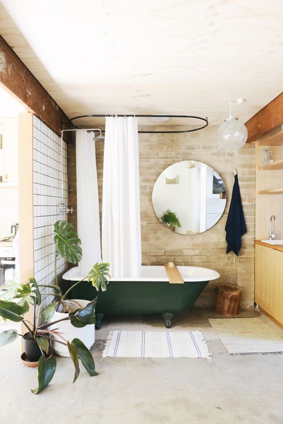 a stylish bathroom with white tiles and brick walls, a dark green clawfoot bathtub, a round mirror, a wooden vanity and potted plants