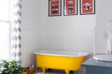 19 a crispy white bathroom with a bold yellow clawfoot bathtub, a blue vanity, a colorful gallery wall and a potted plant