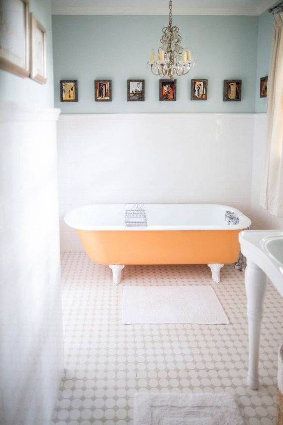 a light-filled bathroom with light blue walls, an orange clawfoot bathtub, a crystal chandelier and a vintage gallery wall