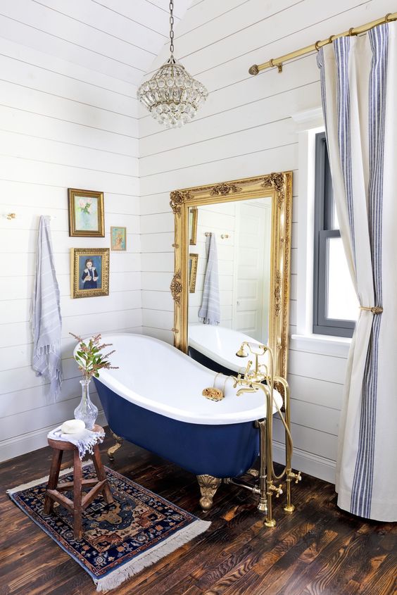 a refined bathroom with white shiplap, a navy clawfoot bathtub, a floor mirror in a gold frame, a crystal chandelier and artworks