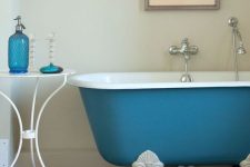 25 a pretty bathroom with neutral walls and a wooden floor, a blue clawfoot bathtub, a side table with blue accessories and an artwork