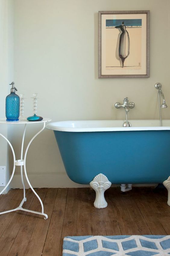a pretty bathroom with neutral walls and a wooden floor, a blue clawfoot bathtub, a side table with blue accessories and an artwork