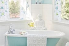 26 a pretty bathroom with light blue walls, a blue clawfoot bathtub, floral print curtains, a white pouf and a crystal chandelier