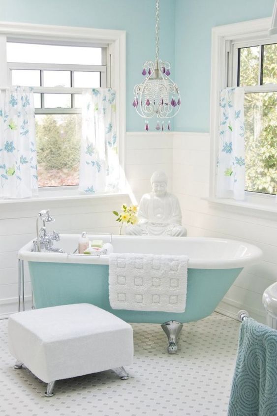 a pretty bathroom with light blue walls, a blue clawfoot bathtub, floral print curtains, a white pouf and a crystal chandelier