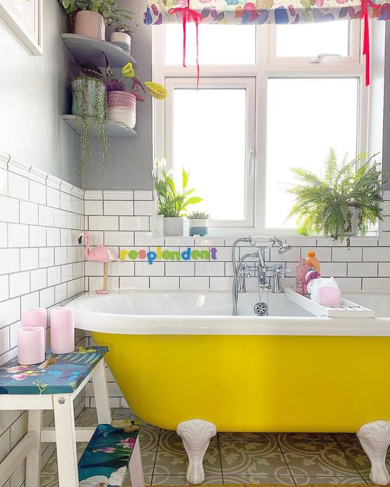 a fun and cheerful bathroom with white subway and tan and white tiles, a yellow clawfoot bathtub, potted plants, touches of pink and bold colors