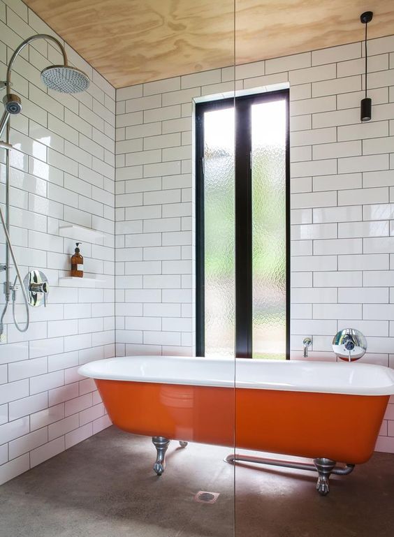 a laconic bathroom with a plywood ceiling, white tiles on the walls, a concrete floor, an orange clawfoot bathtub and a window with frosted glass
